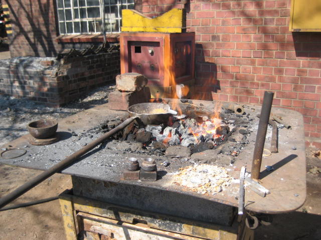 Fanned with compressed air from below, the fire is now hot. The ladle of molten alloy sits on the fire. Two plugs sit at the front of the table.